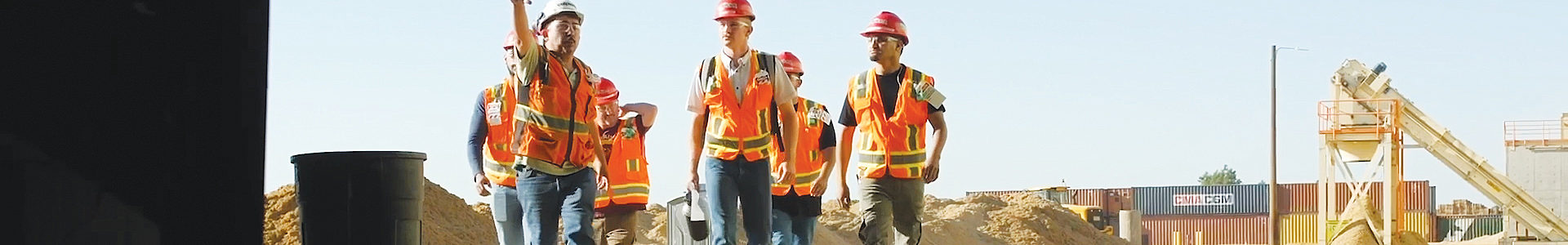 A group of construction workers in conversation walking toward the camera with equipment in the background